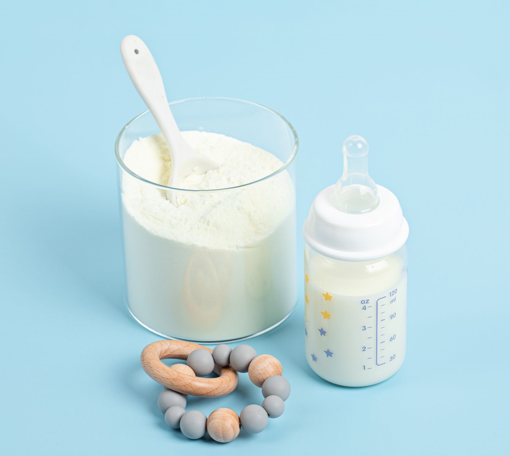 A clear glass filled with high quality baby formula with a white spoon inside it, next to a baby bottle half-filled with formula and marked with measurements. Below the bottle, there is a wooden and silicone teething ring on a light blue background. The items suggest preparation for infant feeding.