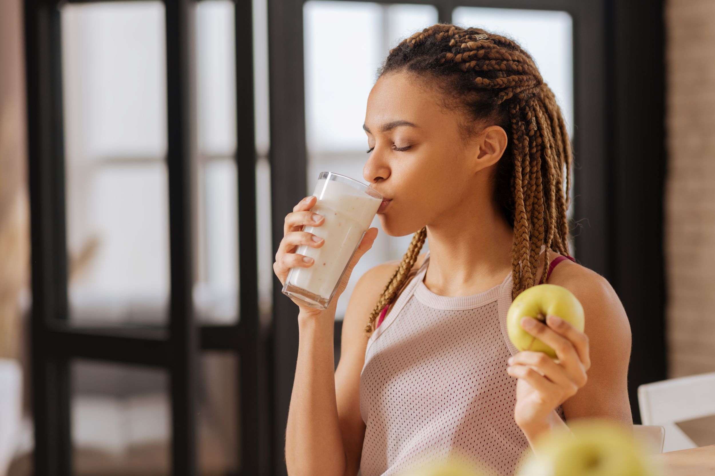 Person with braided hair holding a glass of a high protein dairy shake in one hand and a green apple in the other, standing in a kitchen with a blurred background.