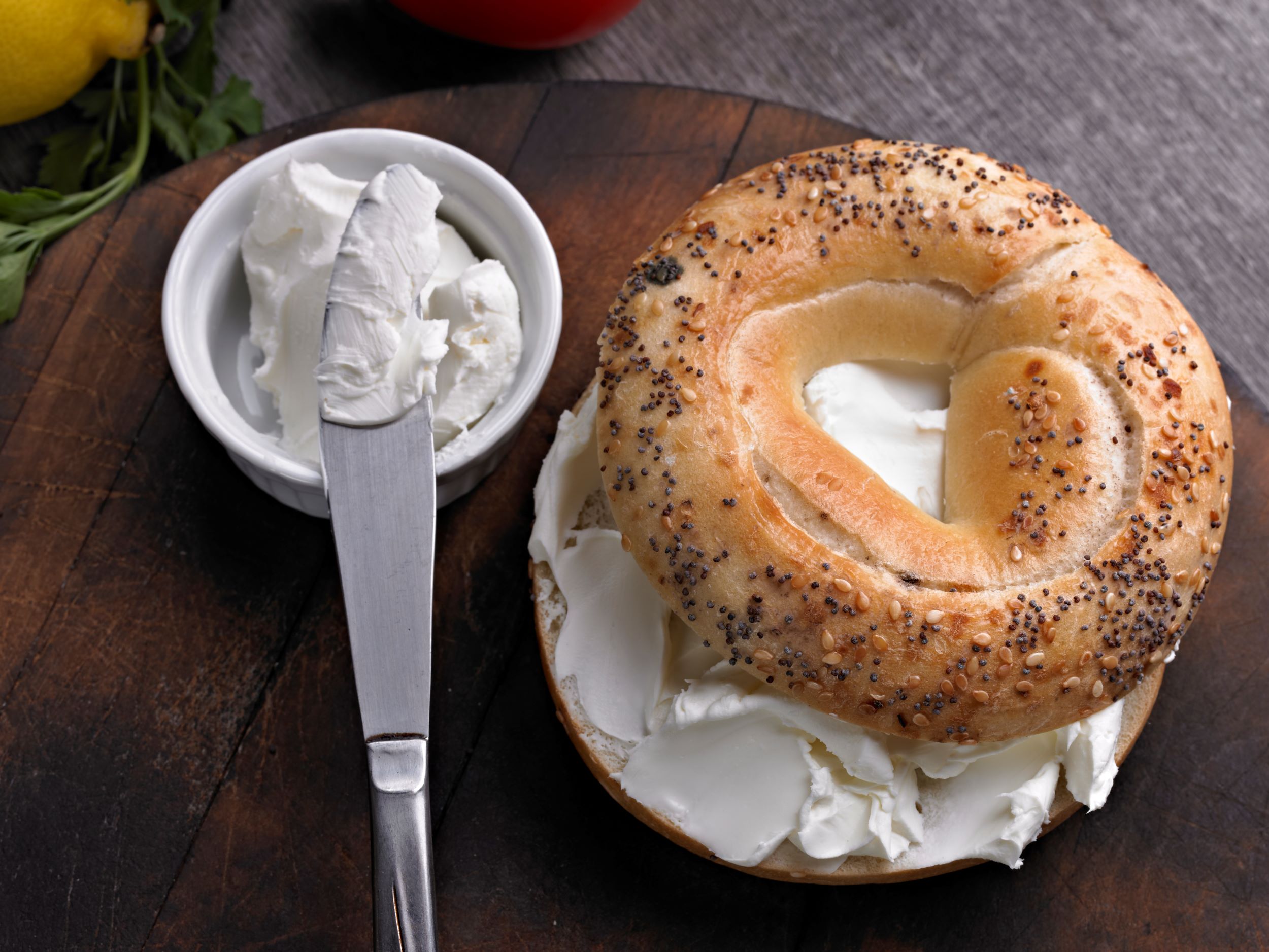 A high protein poppy seed bagel with high protein cream cheese on a wooden surface. One half of the bagel is generously spread with high protein cream cheese, and the other half is placed on top, slightly askew. Next to the bagel is a small bowl of additional cream cheese and a knife with some cream cheese on it. In the background, there are out-of-focus elements including a lemon and some green herbs.