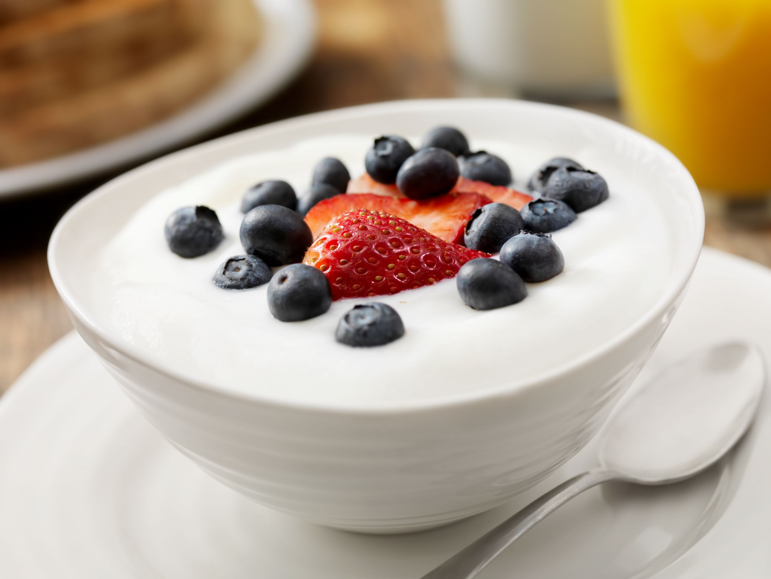 Primer plano de un bol blanco lleno de yogur rico en proteínas con arándanos frescos y una fresa cortada. El bol está colocado sobre una mesa de madera y, al fondo, hay un vaso de zumo de naranja desenfocado y posiblemente más productos para el desayuno. La imagen destaca una opción de desayuno saludable y apetitosa, centrándose en los vibrantes colores y texturas de la fruta frente al cremoso yogur blanco rico en proteínas.