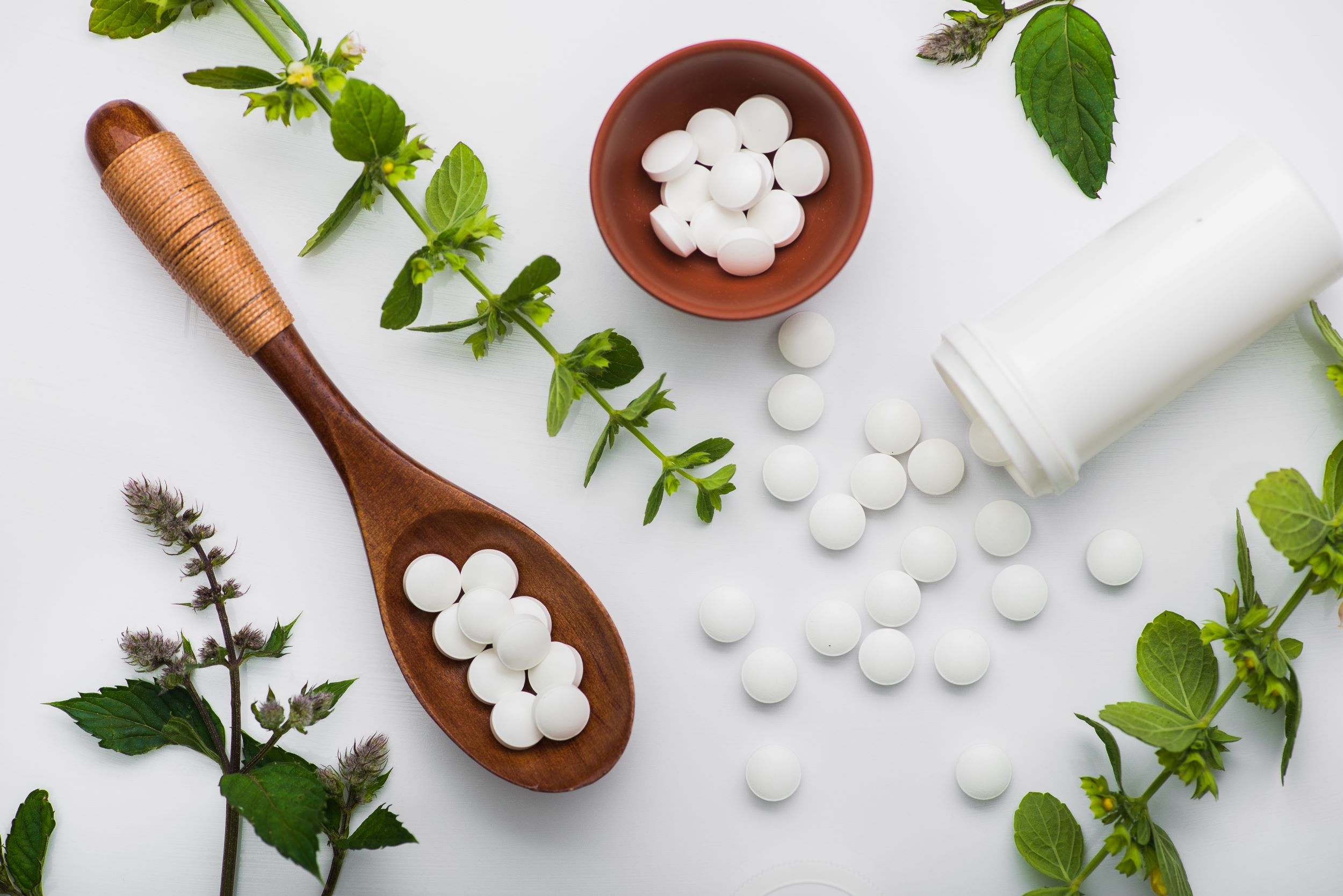 A collection of white nutraceutical tablets some contained within a small brown bowl and others scattered around it on a white surface. To the right, there is an open white pill bottle lying on its side with more tablets spilling out. Surrounding these items are green leaves and herbal flowers, suggesting a theme of natural supplements or medicine.