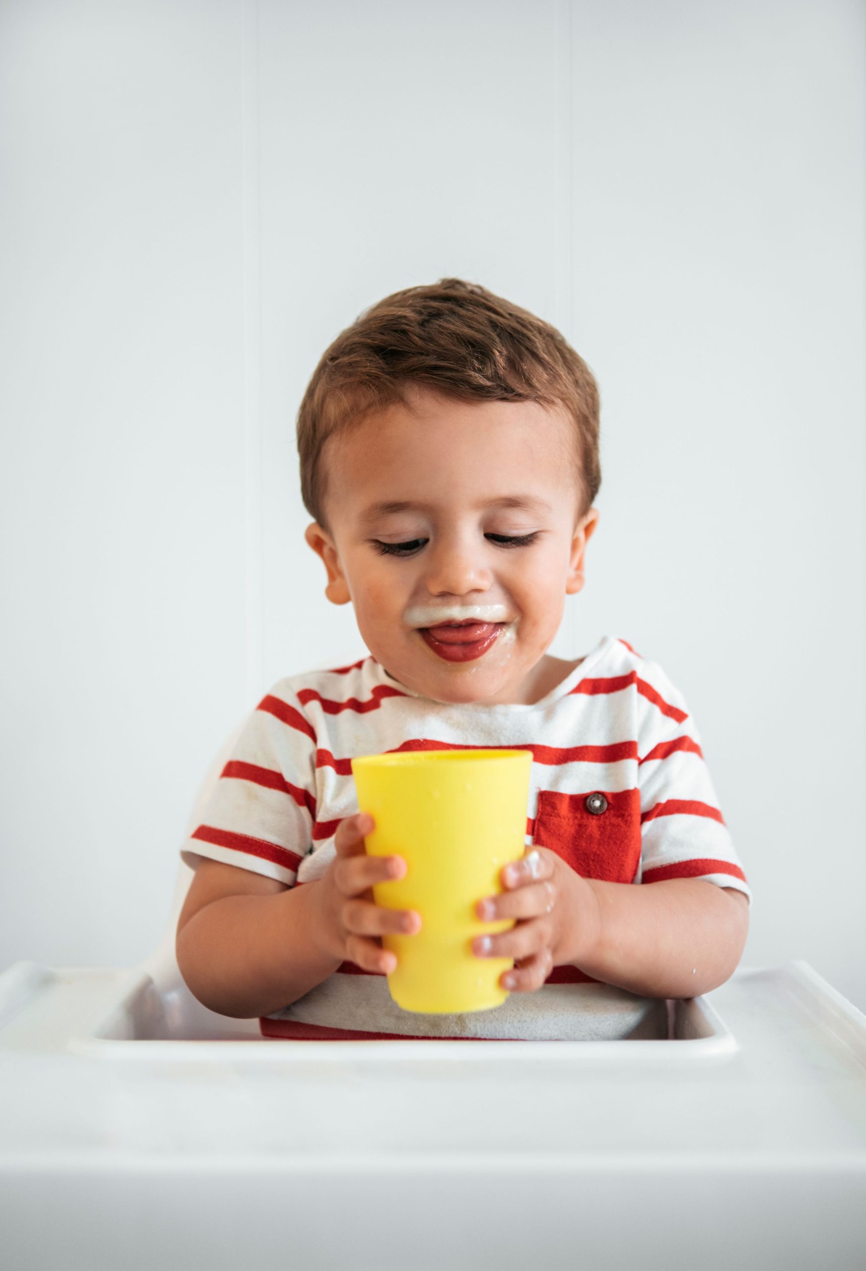 Imagen de un niño pequeño sentado en una mesa, sosteniendo con ambas manos una taza de leche de fórmula proteica para crecimiento. El niño lleva una camiseta a rayas rojas y blancas.