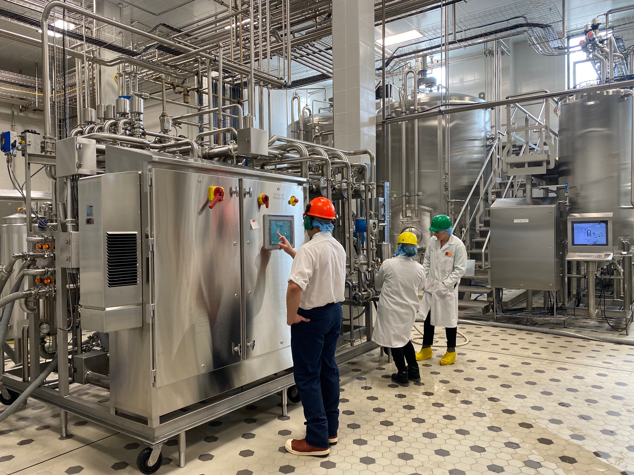 An industrial setting with stainless steel equipment and piping. Four individuals wearing safety helmets, lab coats, and protective footwear are present. One individual is operating a control panel on a large machine, while three others are engaged in a discussion or observation. The floor has a geometric pattern, and there are multiple large cylindrical tanks connected with overhead piping in the background. A digital screen is visible on the right side of the image