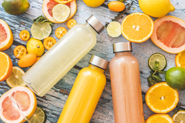 High protein juices lemonade, orange juice, and grapefruit juice in clear bottles with silver caps. Lemons, limes oranges are all scattered in slices and as whole fruit on a wooden table