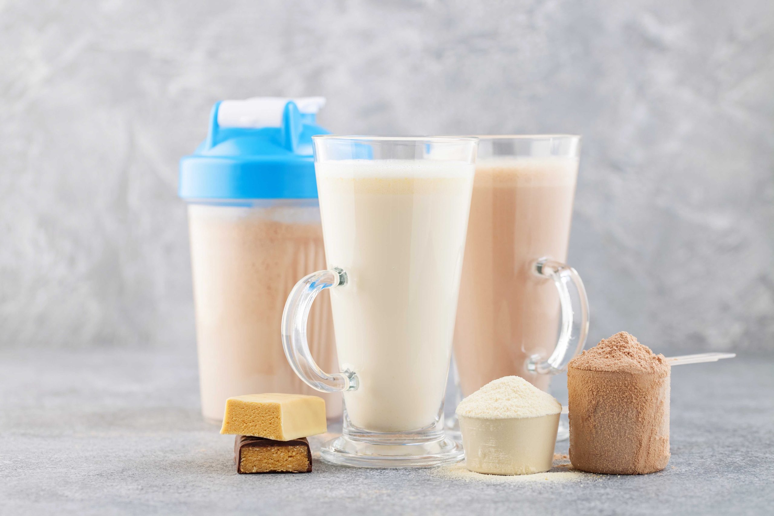 Protein ingredient line up with a vanilla and chocolate protein bars, 1 white powder cup, 1 brown, powder cup, vanilla protein drink and a chocolate protein drink in nice glasses, and a clear protein mixer container with a blue lid