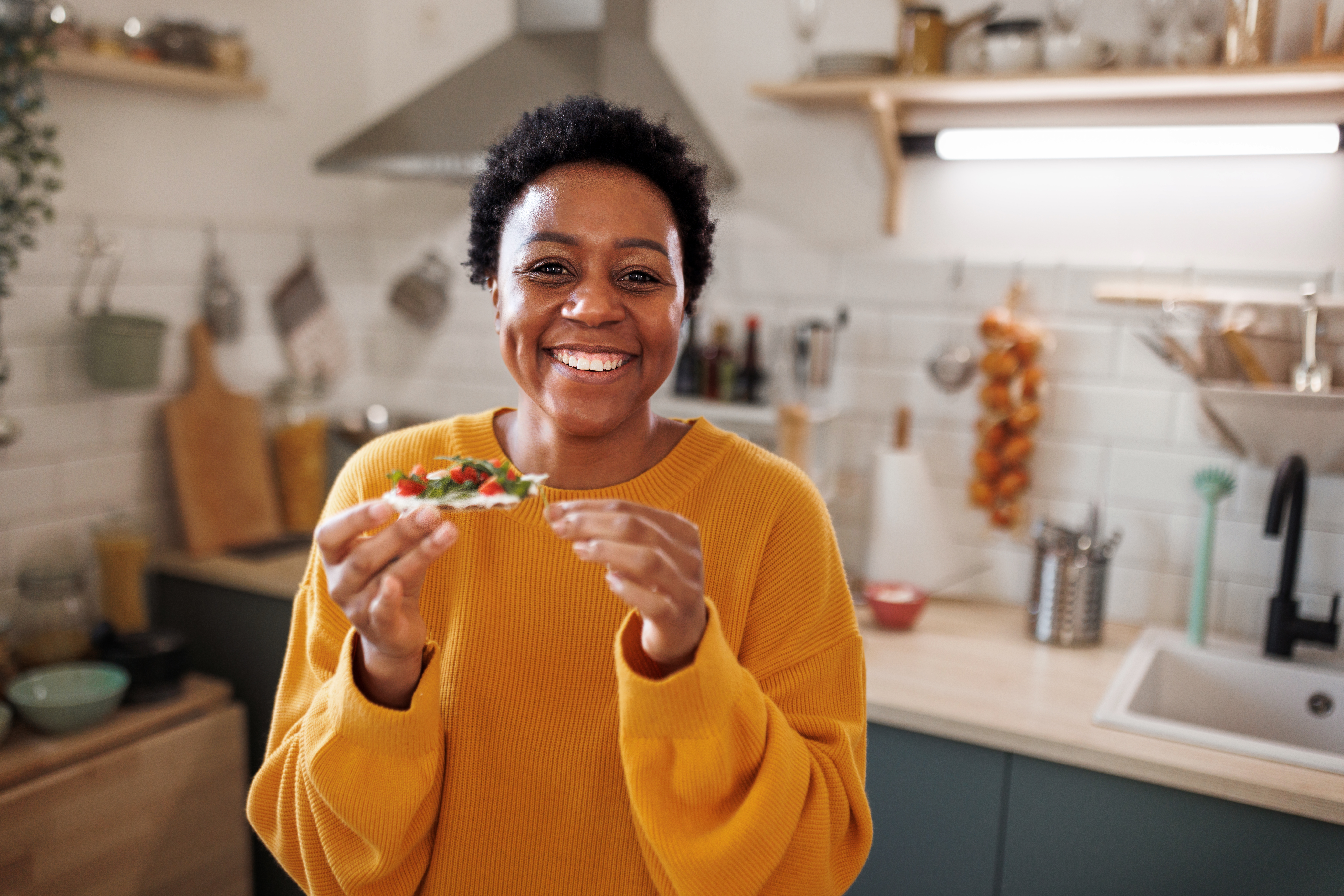 Woman in orange sweater eating a snack and smiling in her kitchen
